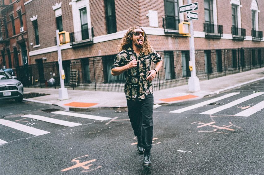 A curly-haired man in sunglasses and a floral shirt runs on a crosswalk in an urban environment with brick buildings and street signs.