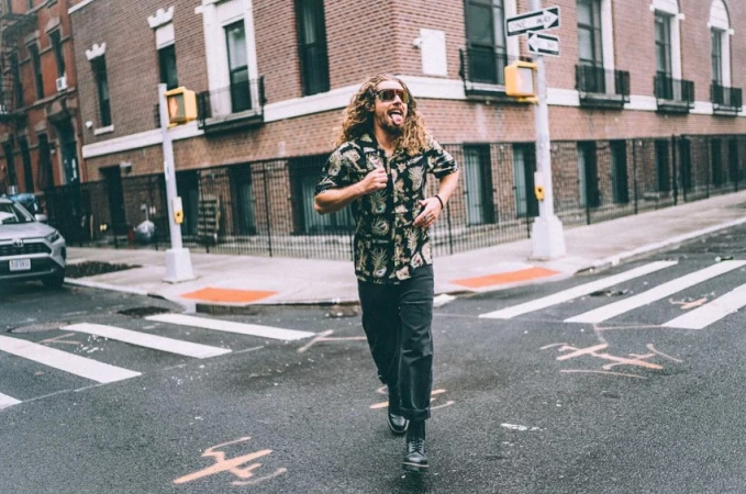 A curly-haired man in sunglasses and a floral shirt runs on a crosswalk in an urban environment with brick buildings and street signs.