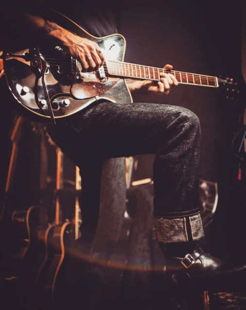 Guitarist playing a shiny electric guitar with black jeans and boots in a dimly lit venue with other instruments nearby.