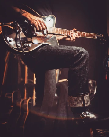 Guitarist playing a shiny electric guitar with black jeans and boots in a dimly lit venue with other instruments nearby.