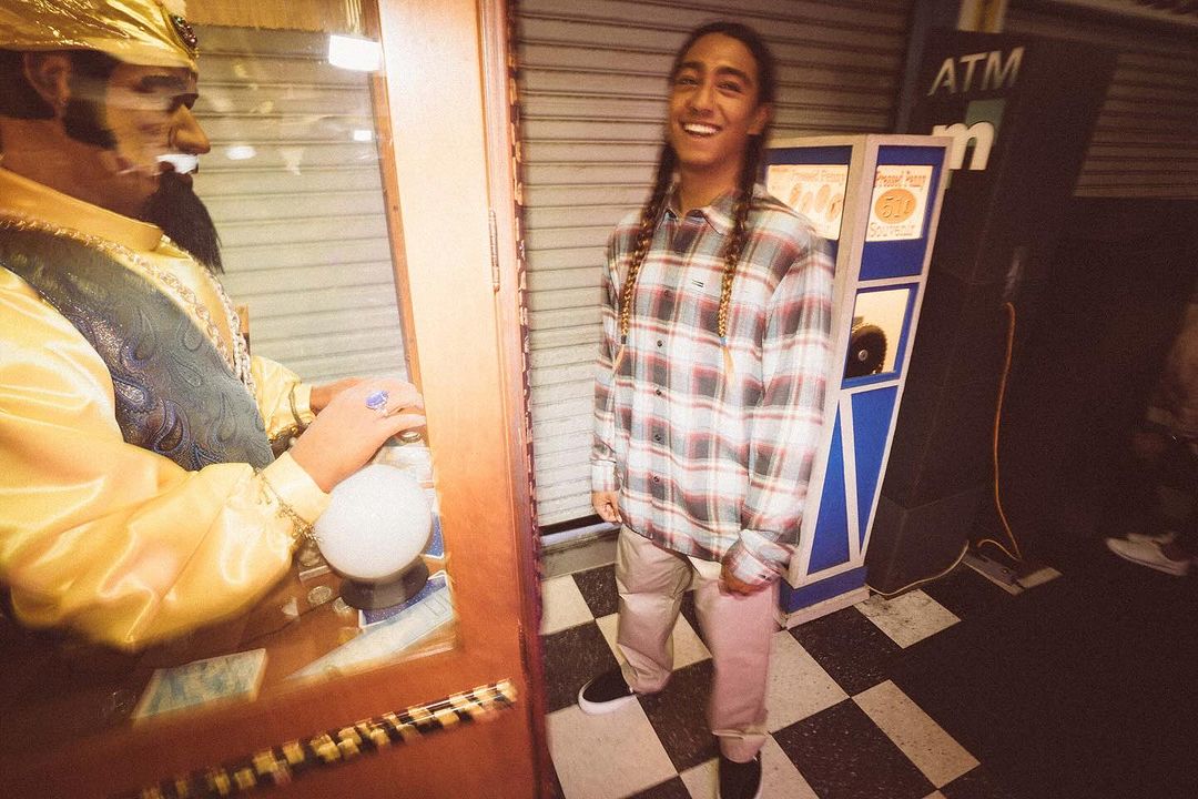 Smiling young man with braided hair wearing flannel shirt and trousers, standing next to a fortune telling machine in an indoor setting.