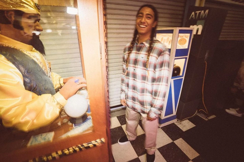 Smiling young man with braided hair wearing flannel shirt and trousers, standing next to a fortune telling machine in an indoor setting.