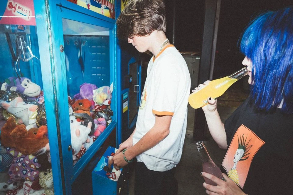 Two young adults, one drinking yellow soda, and another playing a claw machine with plush toys. Casual indoor setting, vintage feel.