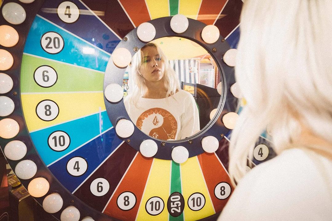 Blonde woman looks in a mirror next to a colorful prize wheel with numbers, inside an arcade setting with vintage color grading.