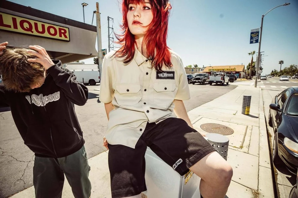 Two young adults wearing Volcom-branded apparel pose on a sidewalk outside a liquor store on a sunny day in an urban environment.