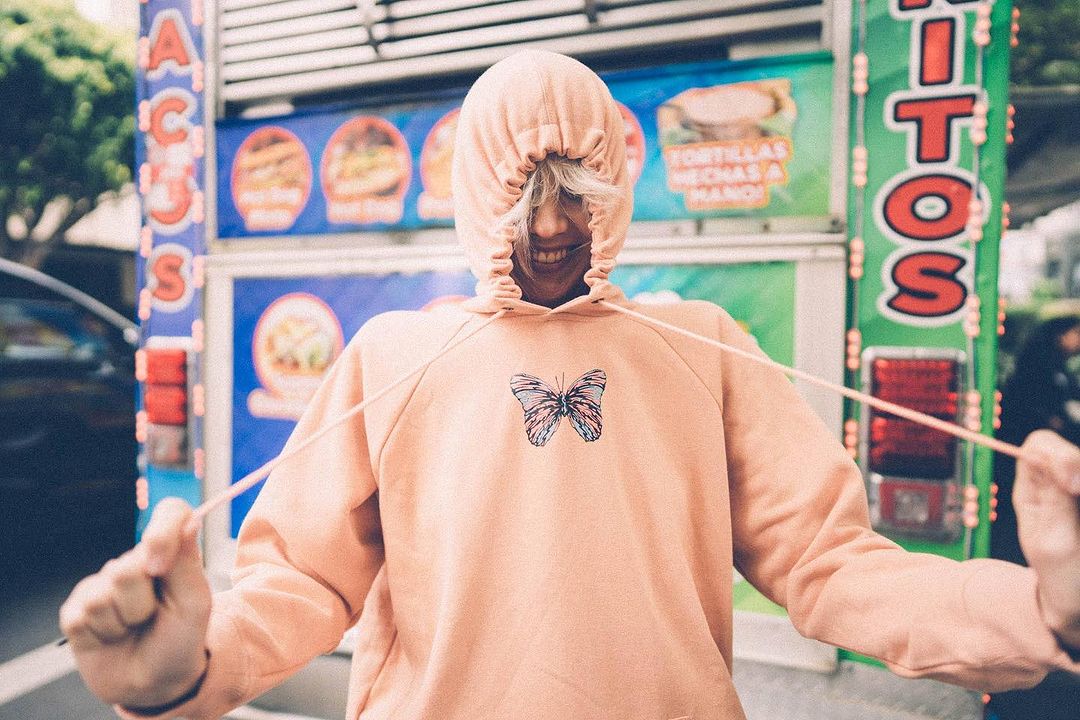 Teen smiles wearing a hooded butterfly sweatshirt standing near a food truck with food illustrations and the words 'Tacos' and 'Tortillas'.