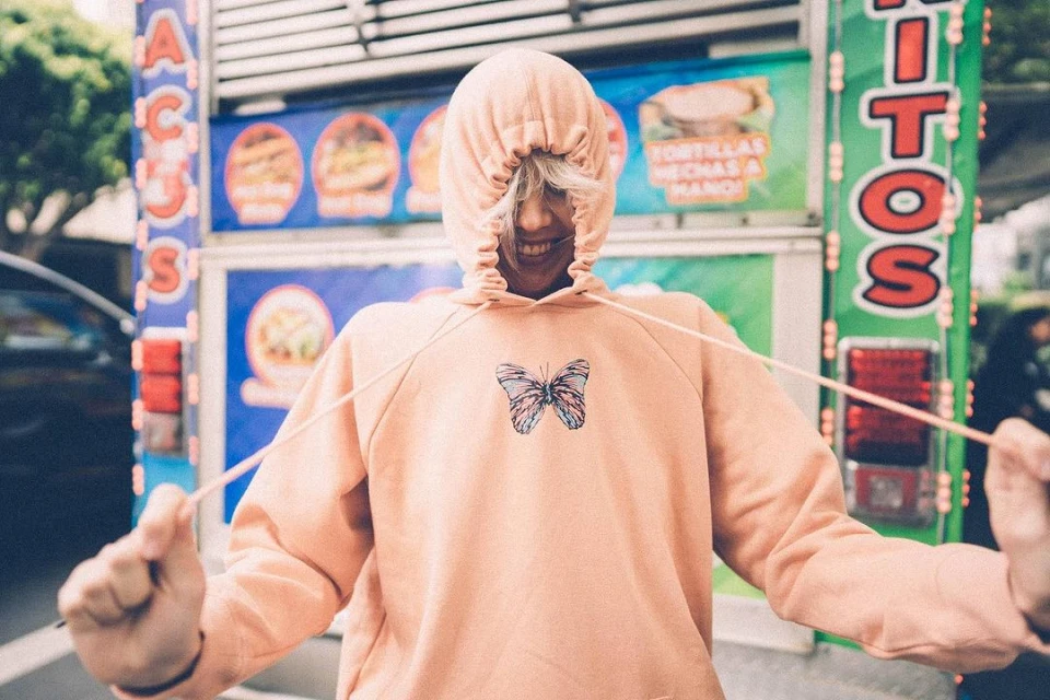 Teen smiles wearing a hooded butterfly sweatshirt standing near a food truck with food illustrations and the words 'Tacos' and 'Tortillas'.