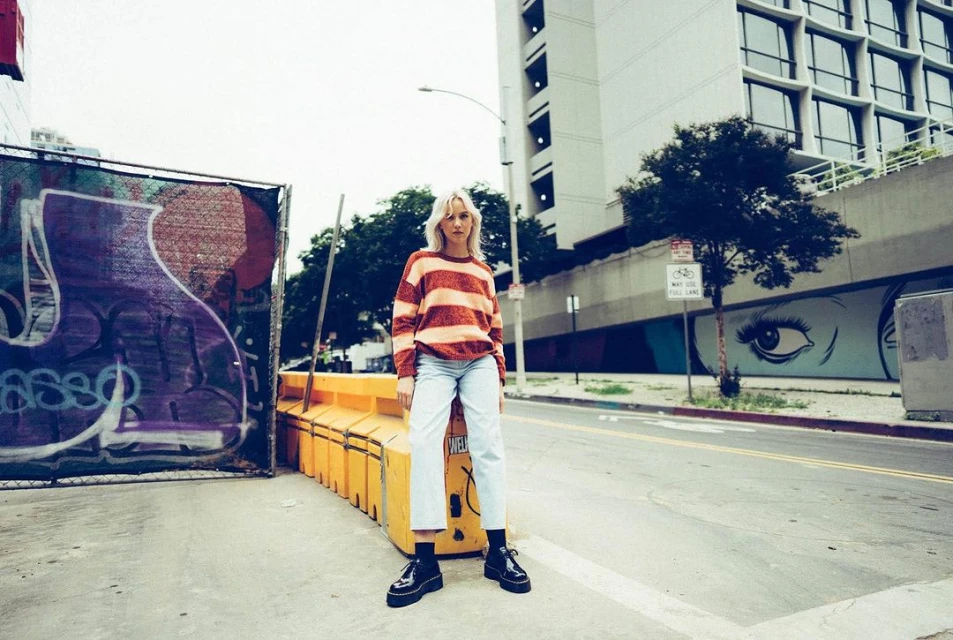 Woman with blonde hair in striped sweater and jeans standing in urban setting in front of building and street graffiti artwork.