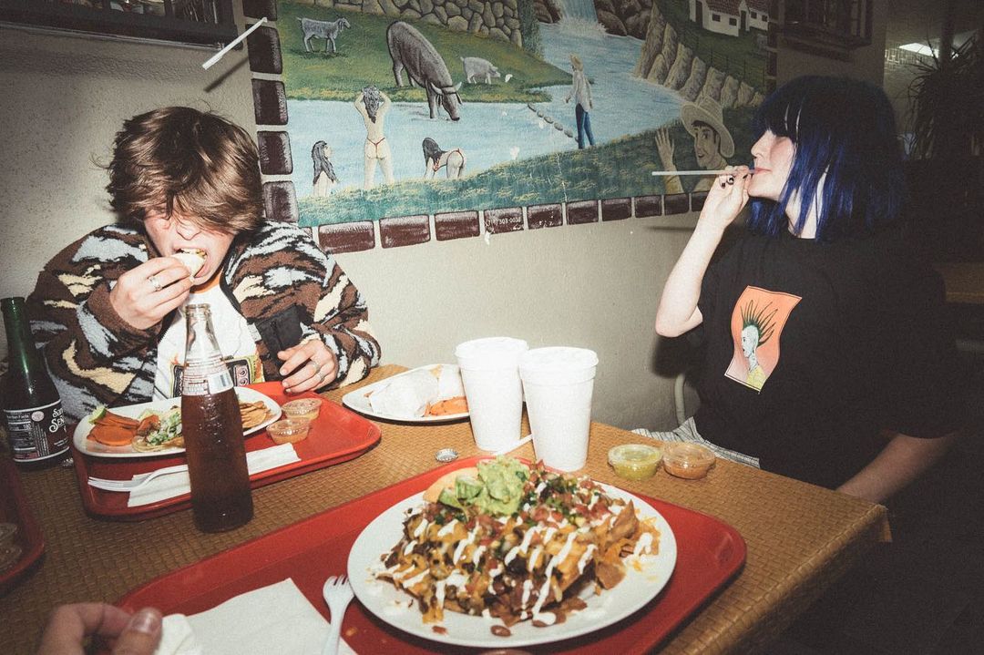 Two people at a restaurant table eating loaded nachos and other Mexican food, sitting in front of a mural with animals and a river.