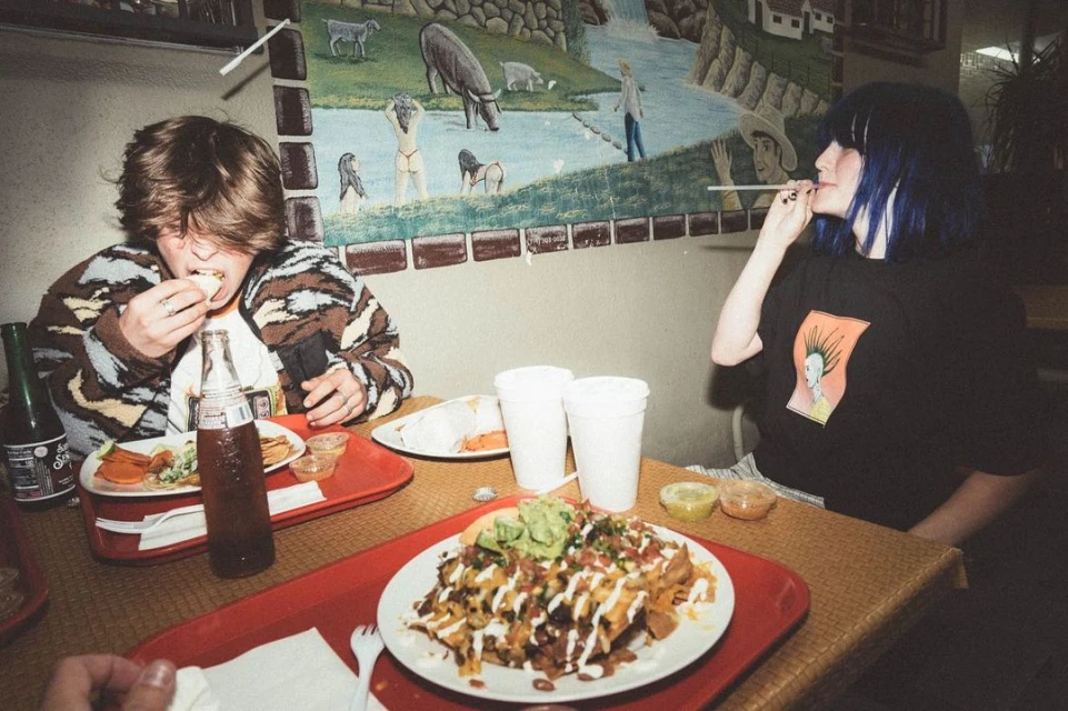 Two people at a restaurant table eating loaded nachos and other Mexican food, sitting in front of a mural with animals and a river.
