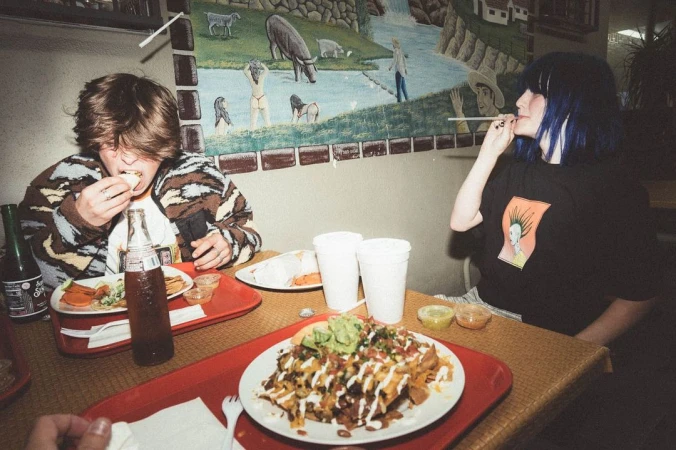 Two people at a restaurant table eating loaded nachos and other Mexican food, sitting in front of a mural with animals and a river.