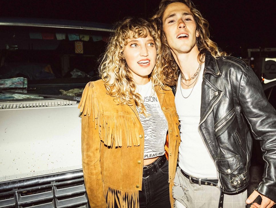 A stylish young couple stands close to a car at night, wearing a brown fringe jacket and a black leather jacket.