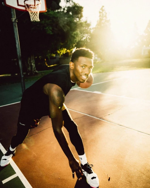 Young Black man dribbling a basketball on an outdoor court with a basketball hoop, wearing athletic apparel, bathed in warm sunlight.