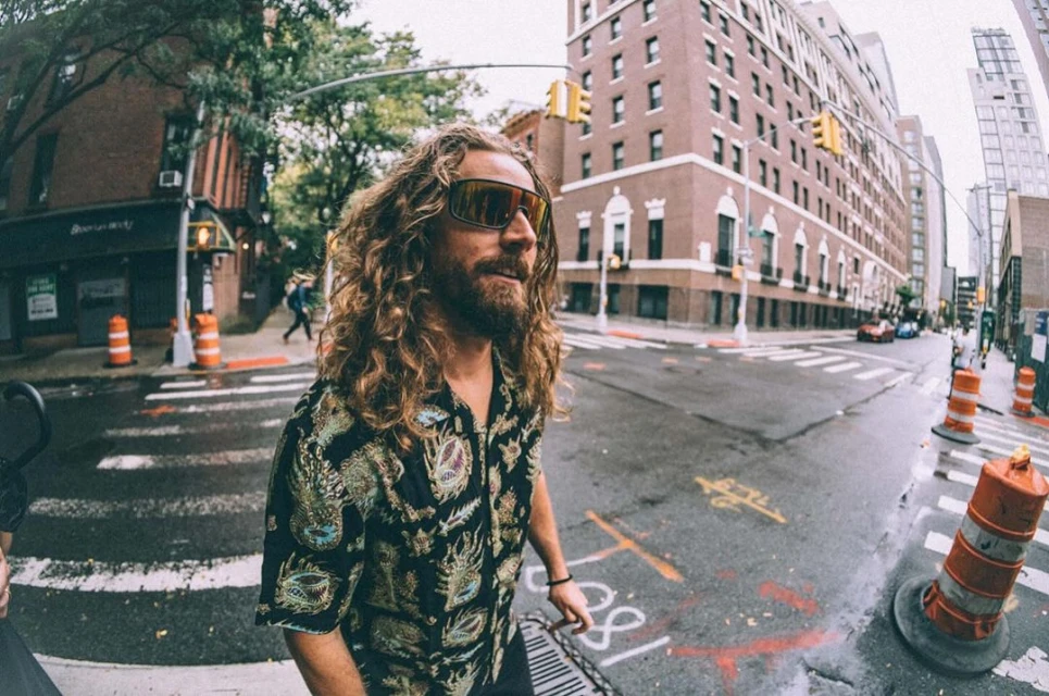 Man with long blond hair and sunglasses stands on a city street wearing a patterned shirt in a wide shot with buildings behind.