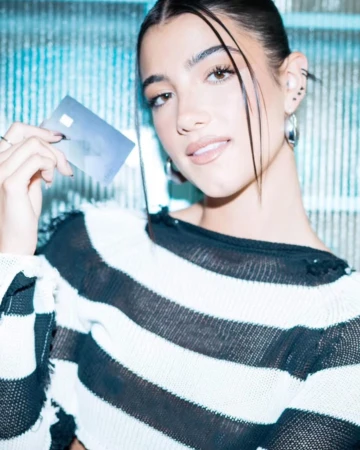 Charli D'Amelio holding a credit card wearing a black and white striped sweater with dark hair tied back.