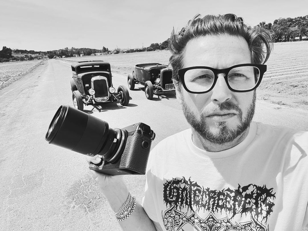 Black and white shot of a man taking a selfie with a camera, with two vintage cars parked on a road in the background on a sunny day.