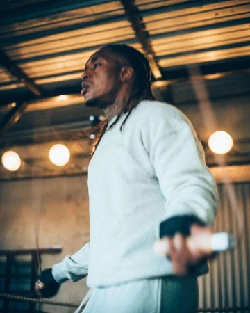 Athletic man in sportswear practices with a jump rope indoors in a gym with industrial ceiling lights and metal boxing ring structure.