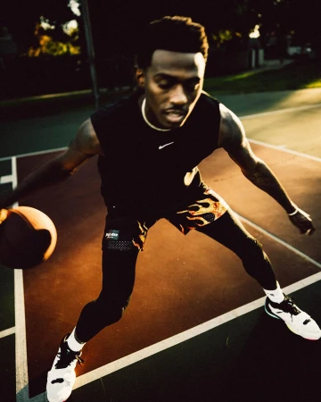 An adult male basketball player dribbles a basketball on an outdoor court, dressed in a black tank top and flame-patterned shorts.