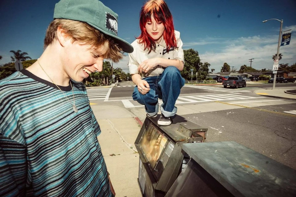 Two young adults, one smiling and one squatting on a metal box, are captured on a city street with a crosswalk and a car in the background.