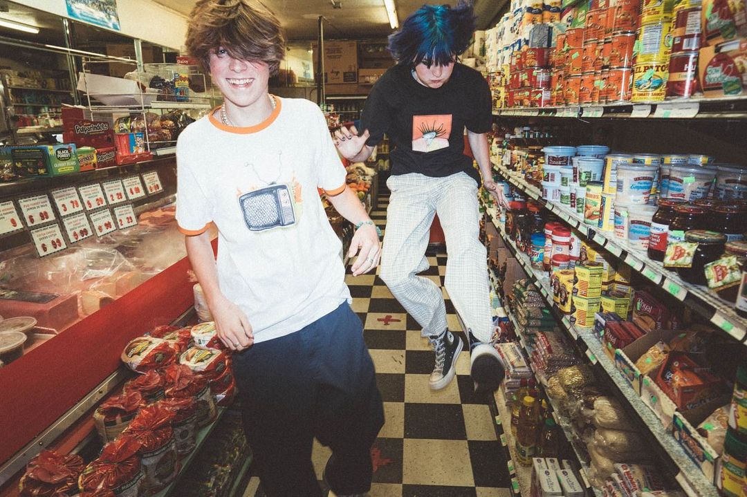Two teenagers in a grocery store aisle, one with blue hair jumping, surrounded by shelves stocked with food products and groceries.