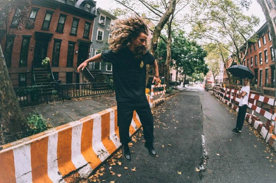 Man with long curly hair leaps mid-air on city street near orange safety barriers while another person stands under umbrella in background.
