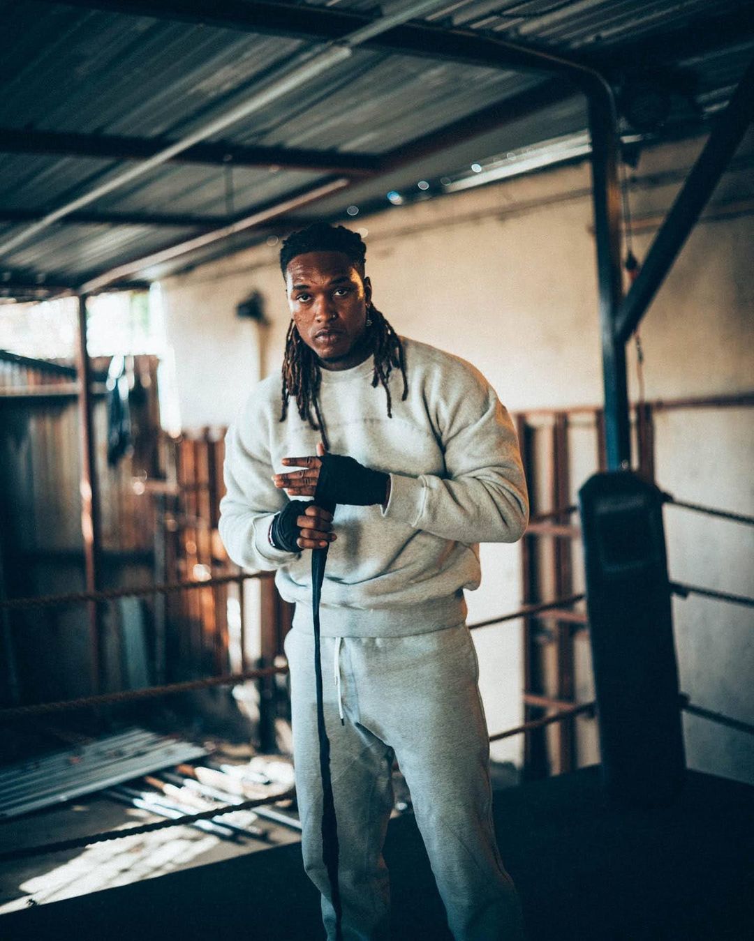 Athletic man with dreadlocks stands in boxing ring wearing grey sweatsuit and hand wraps; ring ropes and metal beams surround.