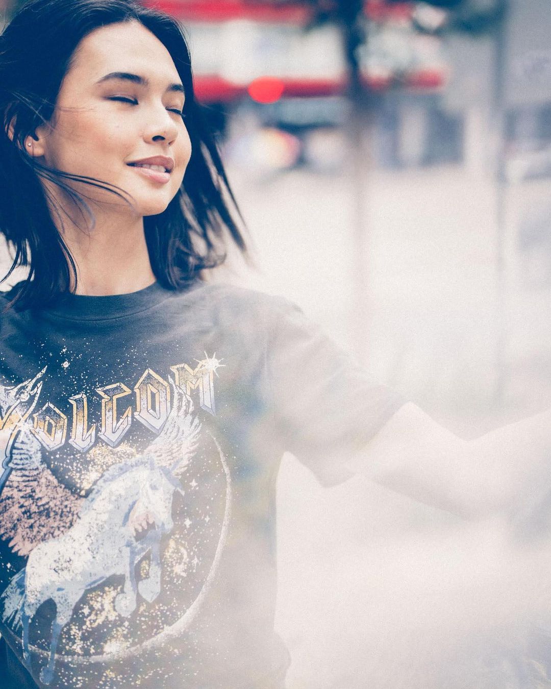 Portrait of a smiling young woman with closed eyes wearing a t-shirt with a unicorn graphic in an out-of-focus outdoor urban setting.