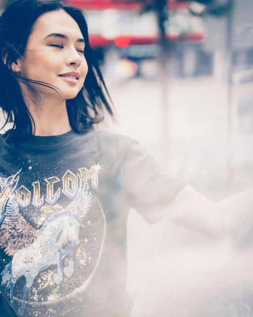 Portrait of a smiling young woman with closed eyes wearing a t-shirt with a unicorn graphic in an out-of-focus outdoor urban setting.