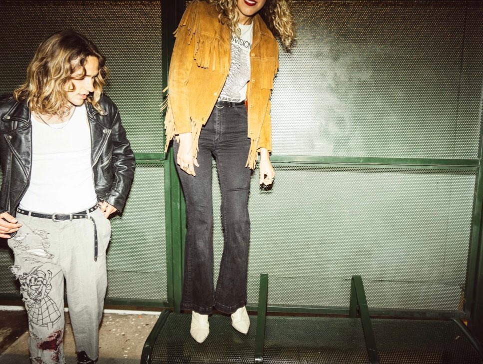 Two people stand by a green perforated wall. One wears a fringed jacket; the other a leather jacket and distressed pants.