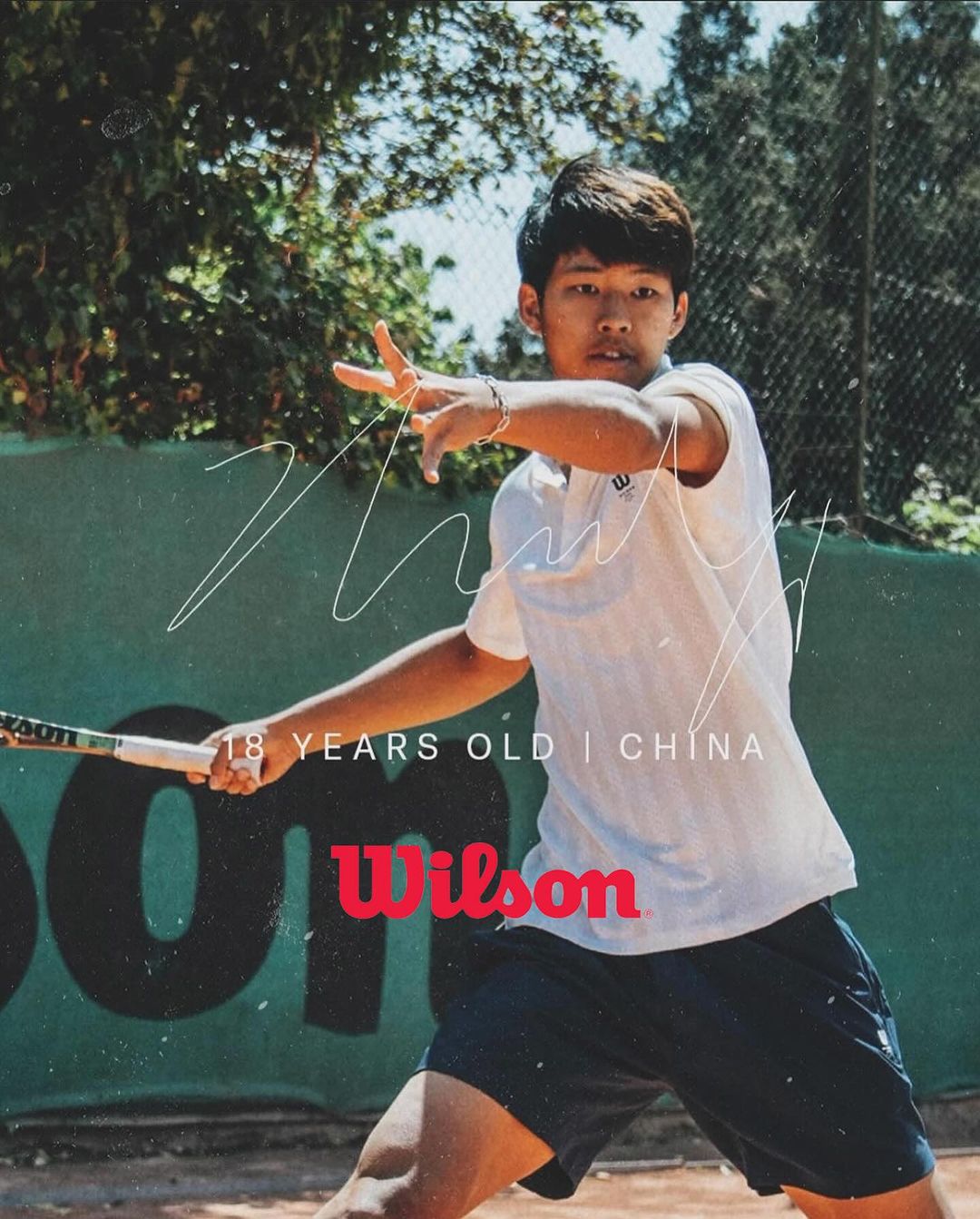 Male tennis player on an outdoor court swinging a Wilson racquet, wearing a white collared shirt and blue shorts, green background.