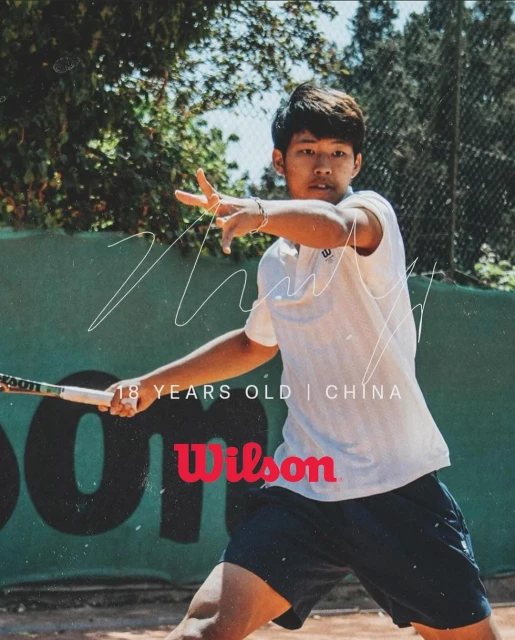 Male tennis player on an outdoor court swinging a Wilson racquet, wearing a white collared shirt and blue shorts, green background.