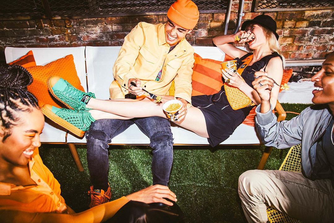 Diverse group of young adults laughing together on an outdoor sofa while sharing food, dressed in casual streetwear with colorful accents.