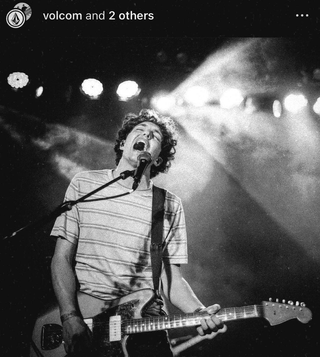 Musician performs on stage, singing into microphone, and playing electric guitar under bright stage lights, in a black and white performance.