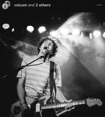 Musician performs on stage, singing into microphone, and playing electric guitar under bright stage lights, in a black and white performance.
