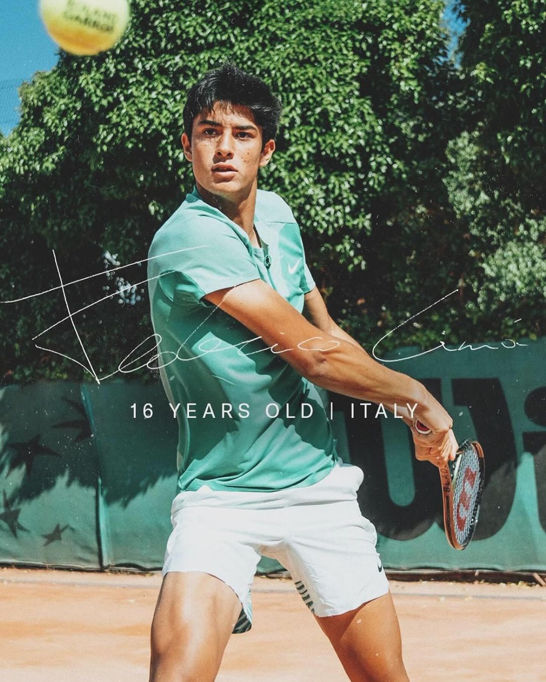 Young tennis player in action with a tennis racket, wearing a green shirt and white shorts, playing on a clay court outdoors.