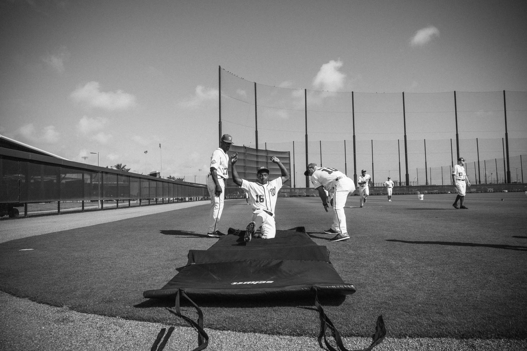 A black and white photograph shows a baseball player kneeling on a mat with arms raised, along with other players and a baseball field.