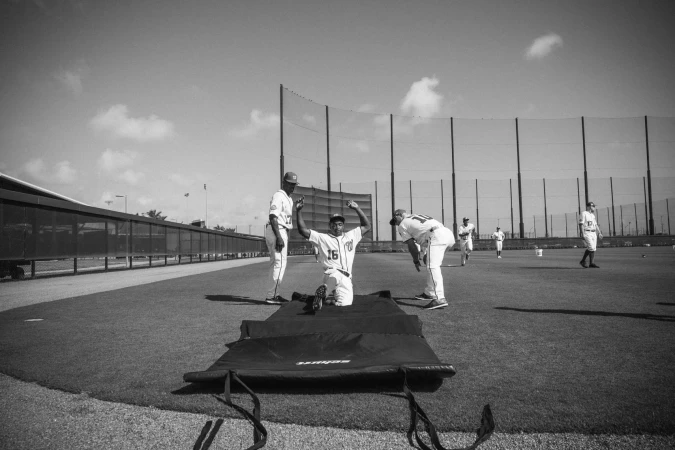A black and white photograph shows a baseball player kneeling on a mat with arms raised, along with other players and a baseball field.