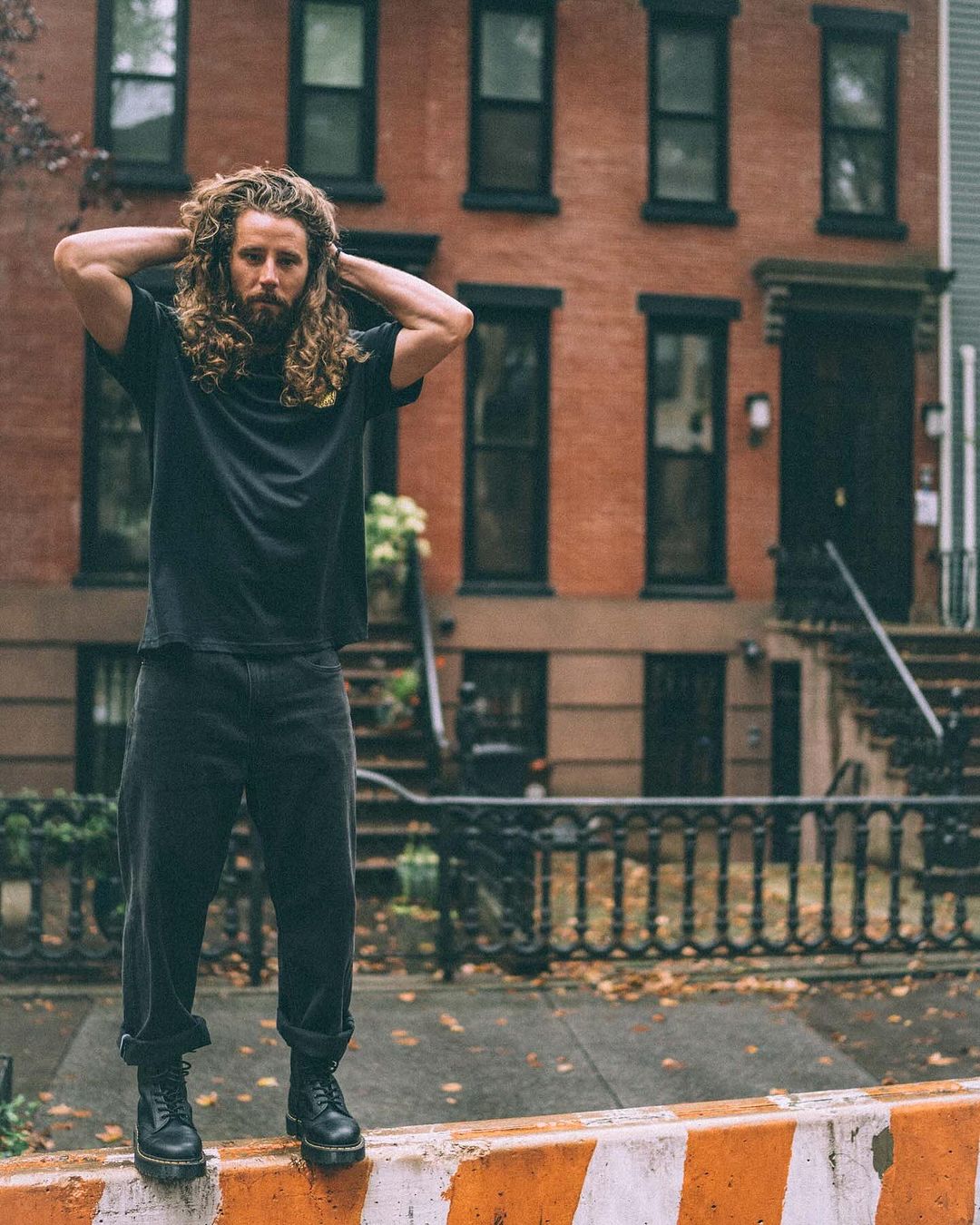 Adult male with long curly hair stands on an orange barrier in an urban setting, wearing a black t-shirt and dark jeans and boots.