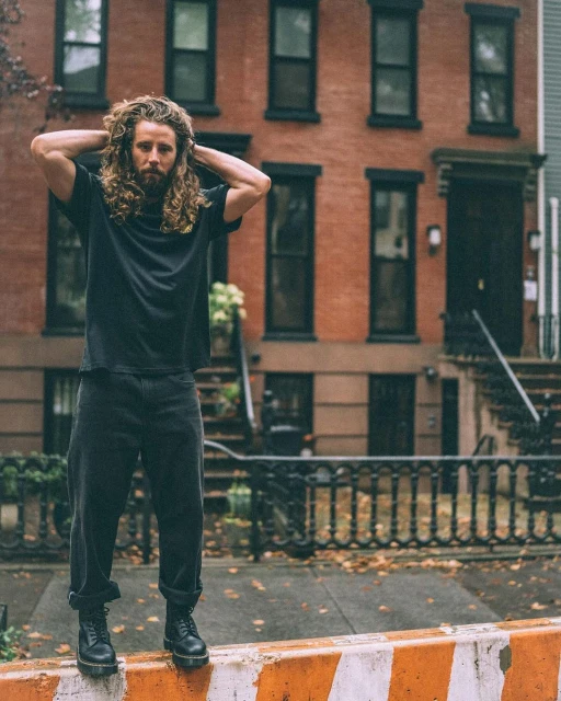 Adult male with long curly hair stands on an orange barrier in an urban setting, wearing a black t-shirt and dark jeans and boots.