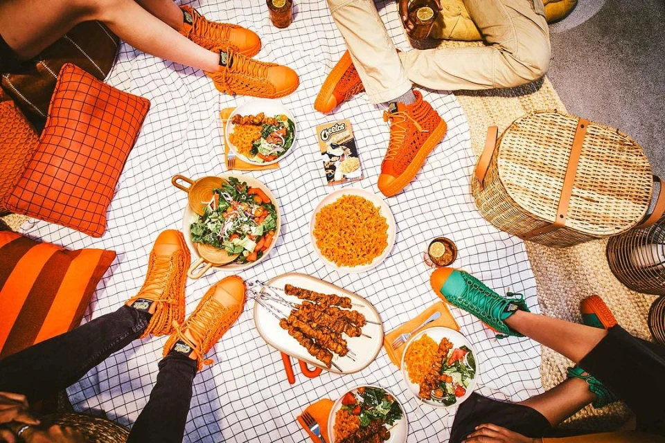 Overhead shot of a picnic blanket with food, drinks, a picnic basket, and people wearing orange and green sneakers, creating a vibrant and inviting scene.