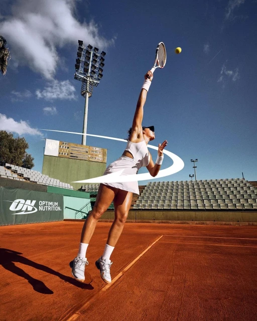 Female tennis player in white sportswear serves the ball on an outdoor court under a blue sky with stadium lighting visible.