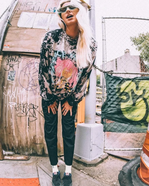 Young woman in patterned top poses in front of graffiti wall in an outdoor urban setting, wearing sunglasses and long blonde hair.