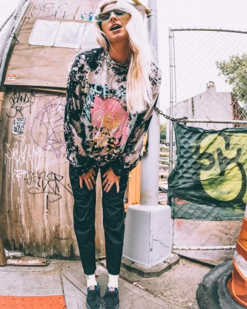 Young woman in patterned top poses in front of graffiti wall in an outdoor urban setting, wearing sunglasses and long blonde hair.