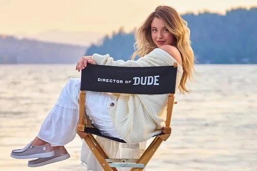A woman with long blonde hair is sitting in a director's chair on the beach near the water at sunset, smiling slightly at the camera.