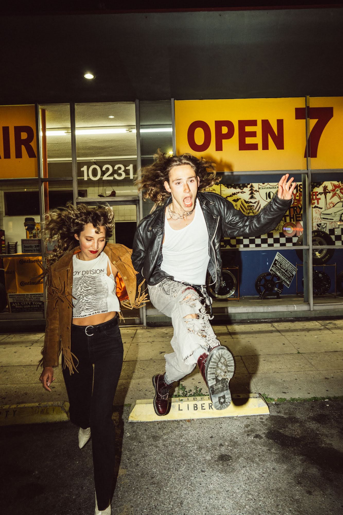 Two young adults, a woman and a man, are outside at night; the man jumps in the air, and the woman walks to the left in front of a building.