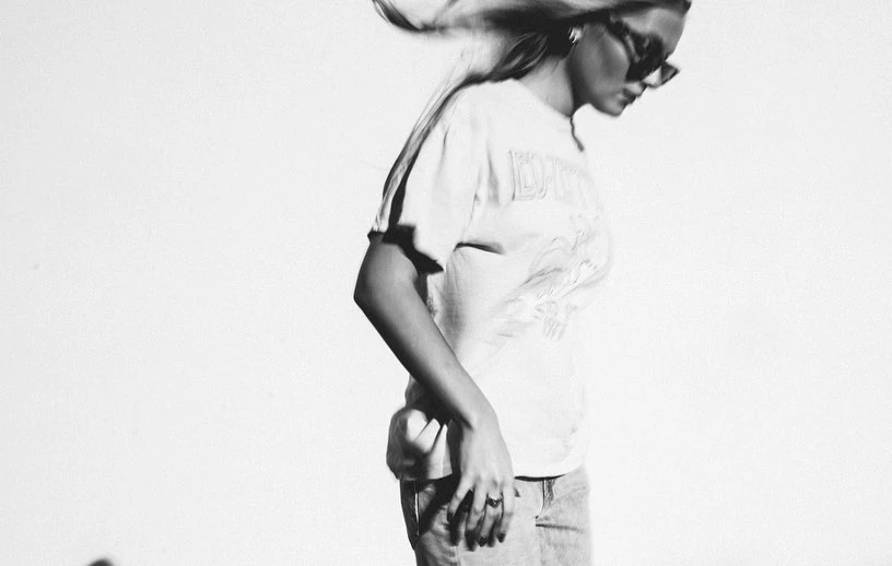 Black and white photo of an adult woman wearing a graphic T-shirt and sunglasses, with her hair blowing in an indoor setting.