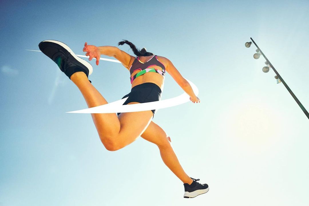 Athletic woman leaps across the finish line with one leg extended high in sportswear against a bright blue sky, athletic shoes.
