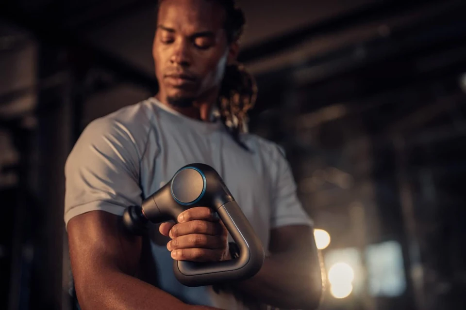 A muscular man uses a massage gun on his upper arm in a dark gym, close up, indoors. Post-workout recovery and muscle therapy for athletes.
