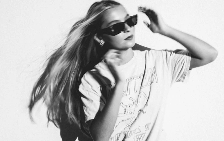 Monochrome portrait of a blond woman wearing dark sunglasses and a graphic t-shirt against a plain white wall