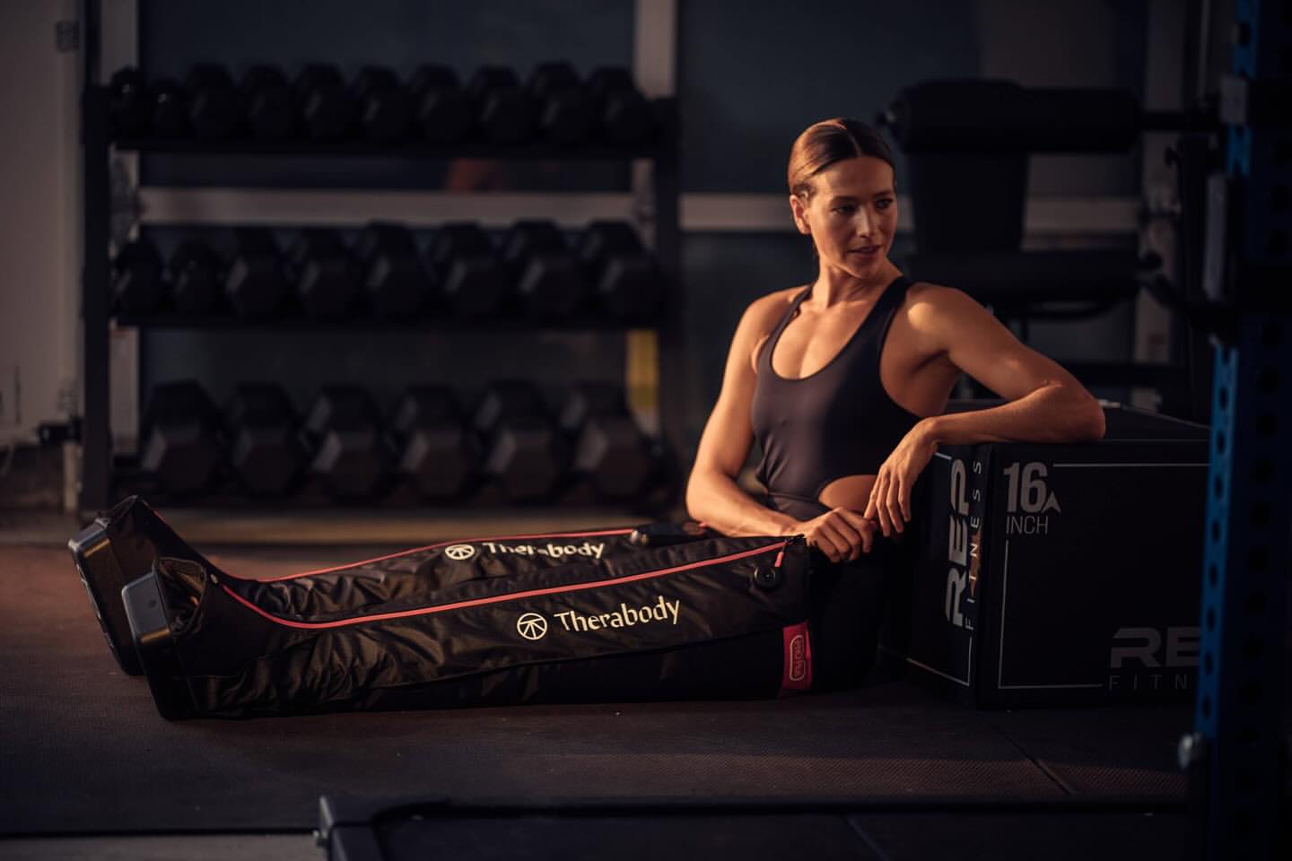 Athletic woman resting with Therabody compression boots in a gym, next to dumbbells. Focus on fitness and recovery.